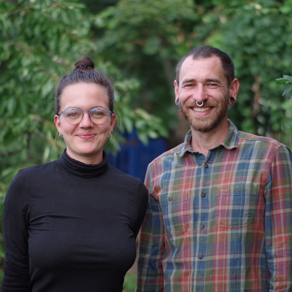 Elisa und Denis stehen vor einem Hintergrund in der Natur. Beide lächeln in die Kamera. 